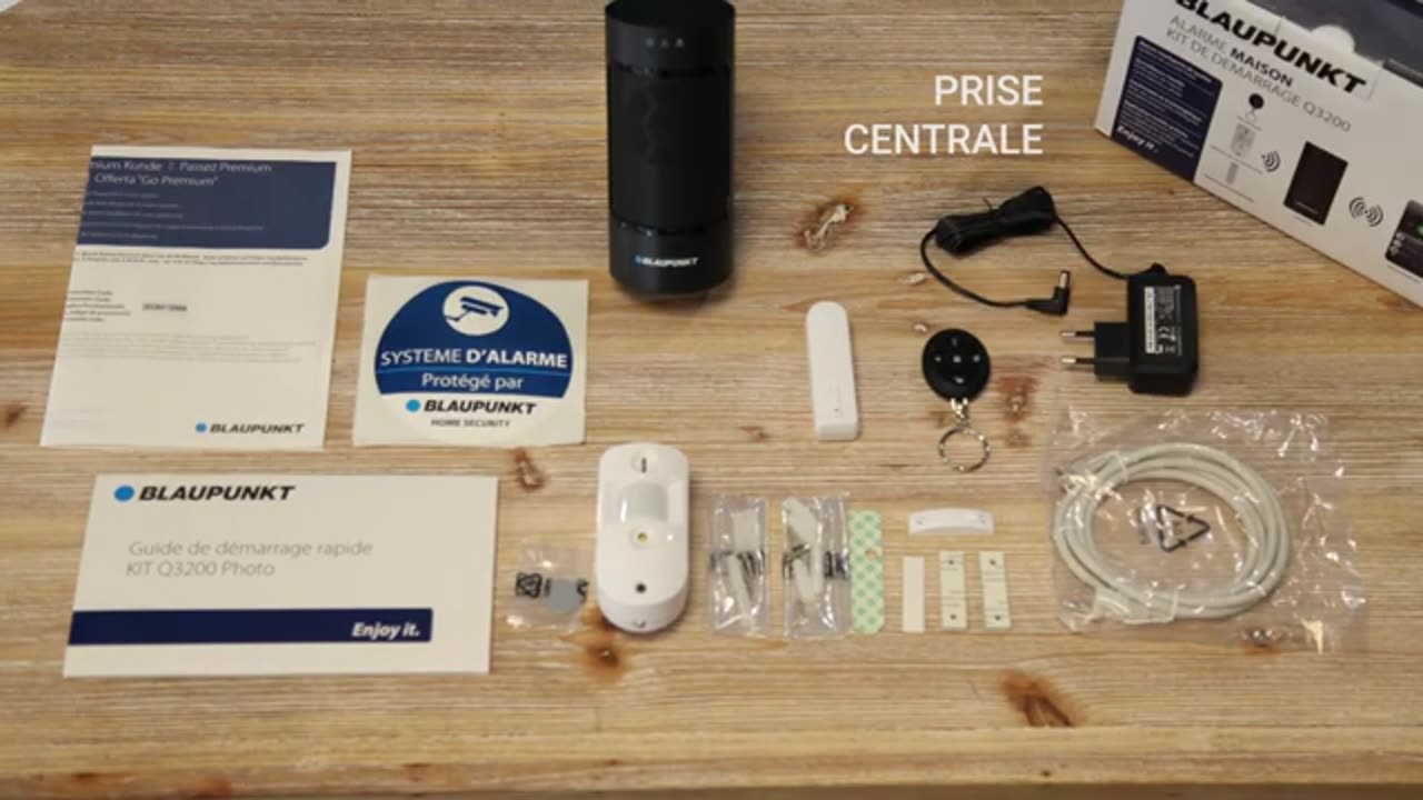 Contenu du kit étalé sur une table : centrale, détecteur, contact, télécommande, câble Ethernet, autocollant et guide
